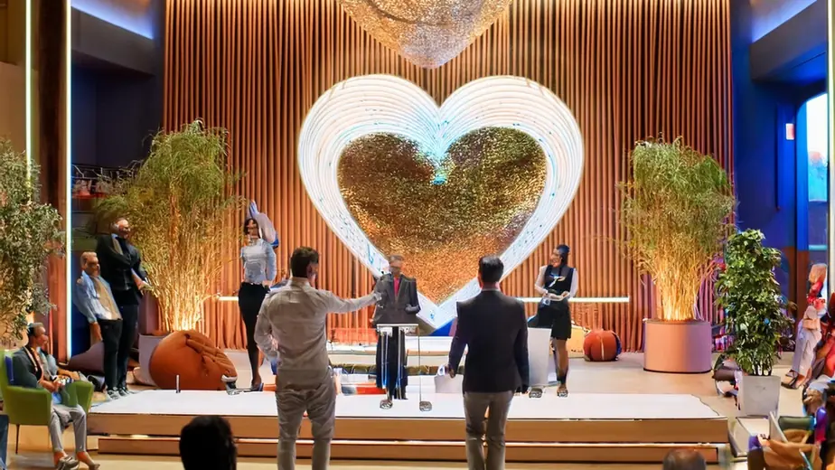 People on a low stage in front of a large illuminated heart, watched by a seated audience