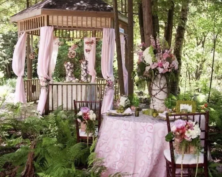 A wooden gazebo draped in pink fabric with a table laid for a meal, surrounded by ferns