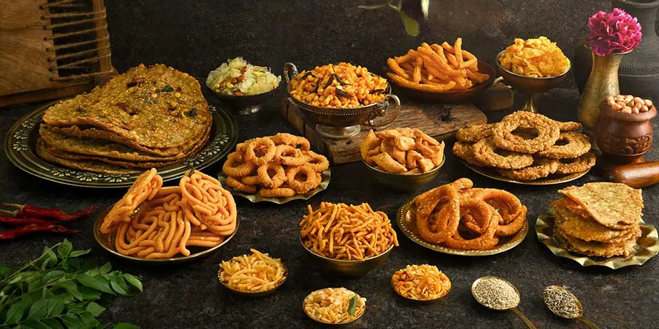 Brass plates of Indian fried snacks including chakli, sev and murukku on a dark table